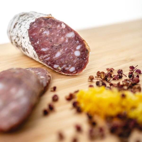 A close up picture of a cross section of lemongrass szechuan peppercorn salami with herbs and spices in the foreground.