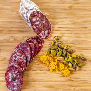 A salami called Orange Pistachio salami pictured from above on a cutting board. One half of the salami chub is cut into slices while the other half remains whole next to pistachios and orange zest.