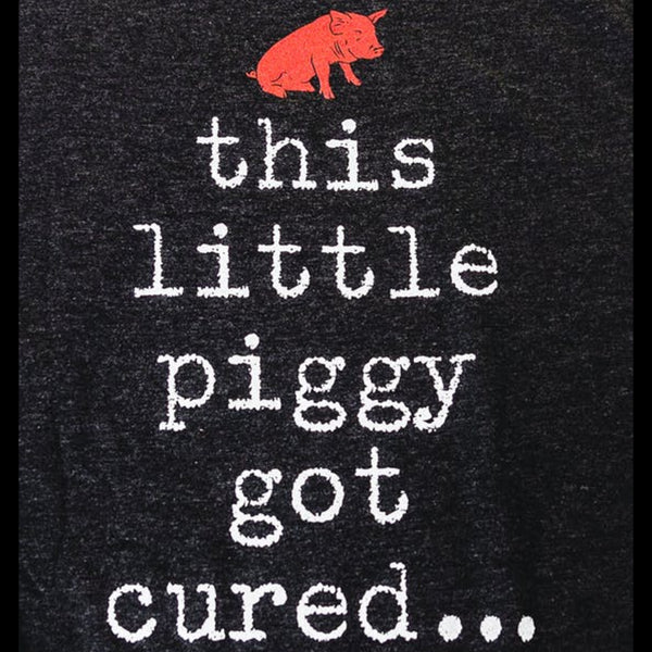 A close up of il porcellino salumi's pig logo and the words "this little piggy got cured..." screen printed on a black shirt.
