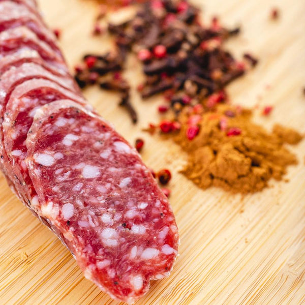A close up of salami slices with fat, herbs and spices detailed in the salami. The slices are on a cutting board with blurred herbs and spices to the right of it.