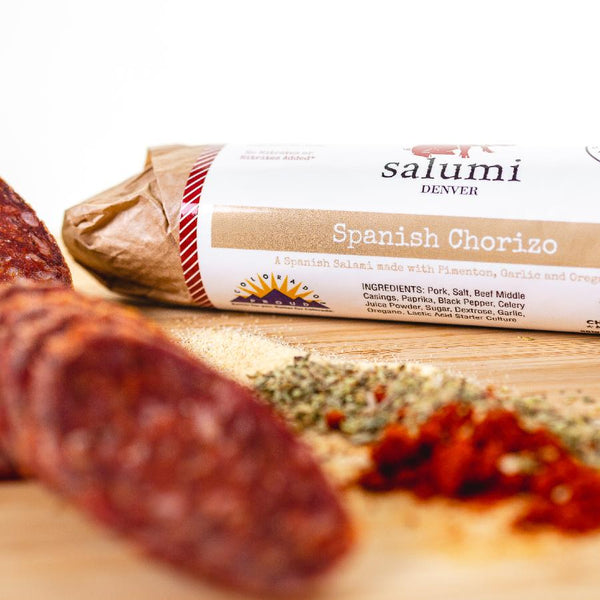 A close up of Spanish Chorizo salami in packaging on a cutting board. There are salami slices, herbs and spices blurred in the foreground.