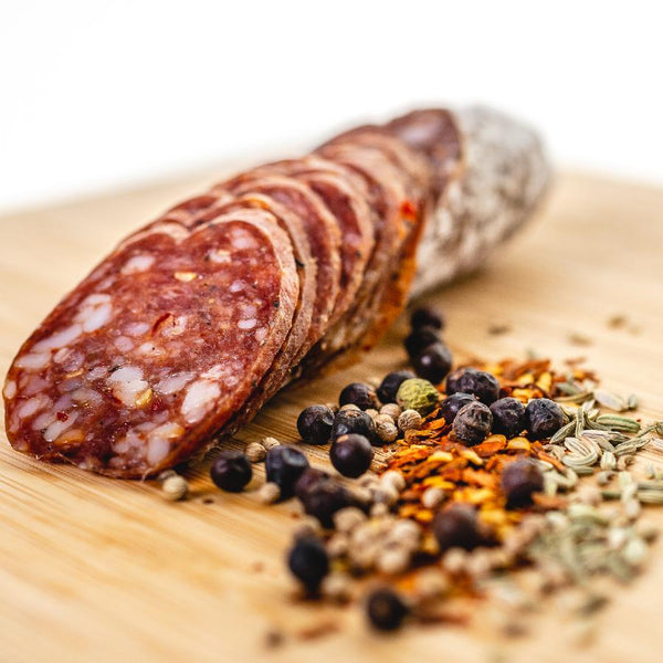 A chub of salami on a cutting board with half of it cut into bite size slices. Juniper berries and other herbs and spices are in the right foreground.