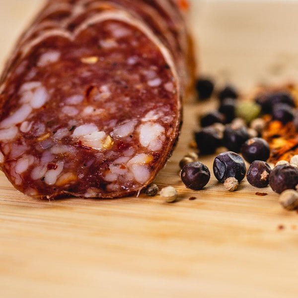 A photo looking directly at a slice of salami taken close up so you can see the fat, herbs and spices. The salami slices are on a wood cutting board with juniper berries and other spices to the right of them. 