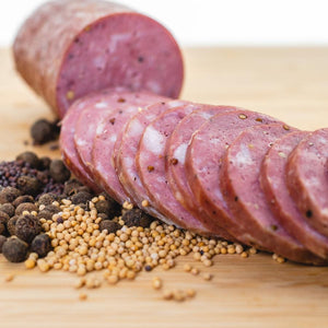 A chub of summer sausage on a cutting board with half cut into edible slices. The uncut half is in the background and herbs and spices are in the foreground.