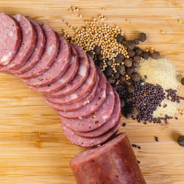 A bunch of summer sausage slices and a whole chub of summer sausage on a cutting board next to herbs and spices and pictured from above.